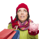 Stressed Mixed Race Woman Wearing Winter Clothing Looking Up Holding Shopping Bags and Piggybank Isolated on White Background.